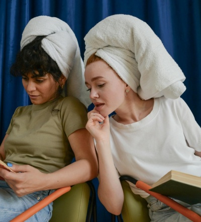 Two women with their hair up in towels. They are reading books sitting in a chair, one in white t-shirt is looking into the green t-shirt woman's book.