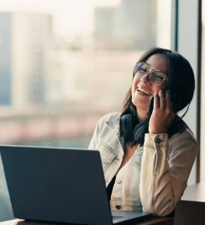 Woman laughing while talking in her phone. She is in her office and has a laptop open.