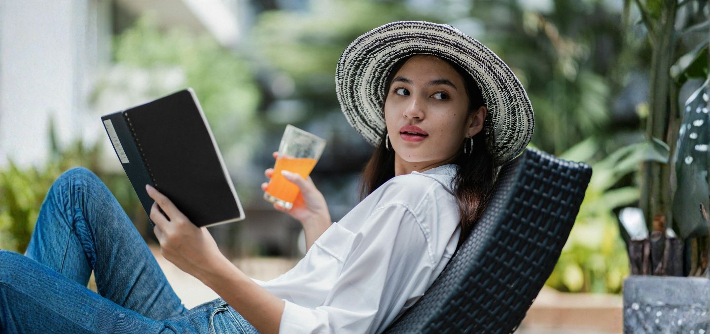 woman reading a book with a orange drink in her hand. She's wearing a vacation hat.