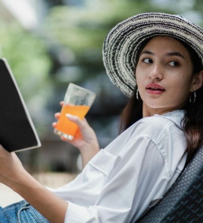woman reading a book with a orange drink in her hand. She's wearing a vacation hat.